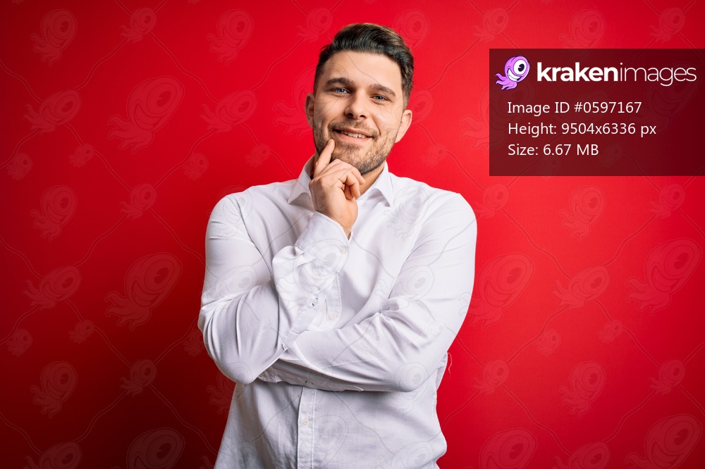 Young business man with blue eyes wearing elegant shirt standing over red isolated background looking confident at the camera smiling with crossed arms and hand raised on chin. Thinking positive.