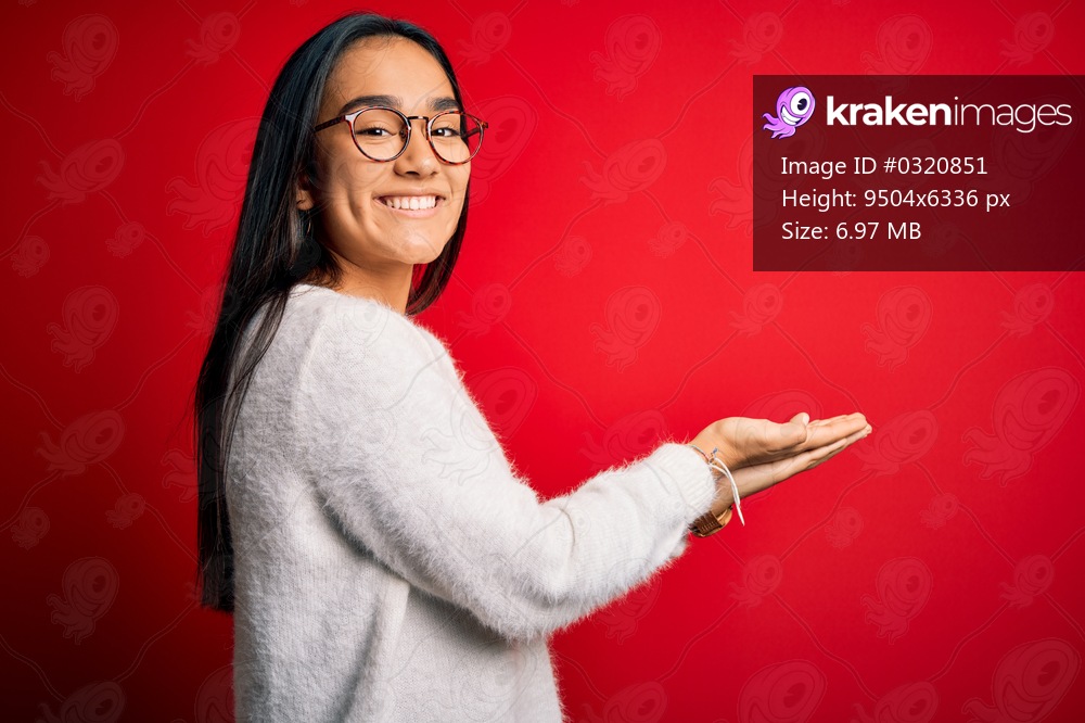 Young beautiful asian woman wearing casual sweater and glasses over red background pointing aside with hands open palms showing copy space, presenting advertisement smiling excited happy