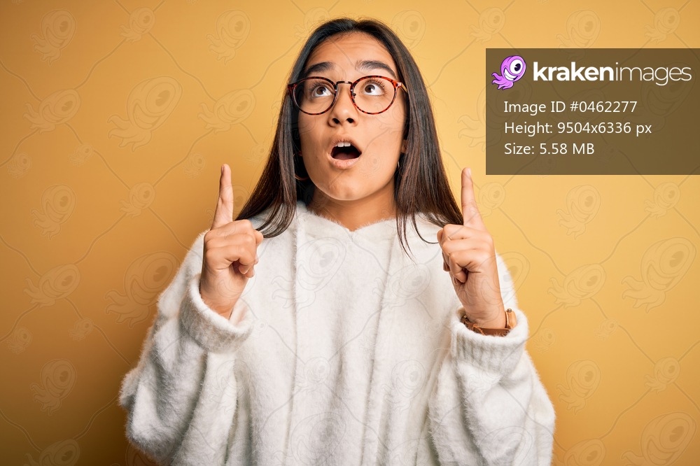 Young beautiful asian woman wearing casual sweater and glasses over yellow background amazed and surprised looking up and pointing with fingers and raised arms.