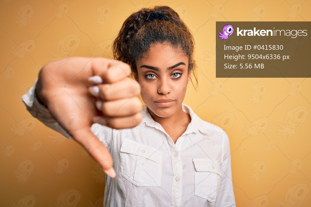 Young beautiful african american girl wearing casual shirt standing over yellow background looking unhappy and angry showing rejection and negative with thumbs down gesture. Bad expression.