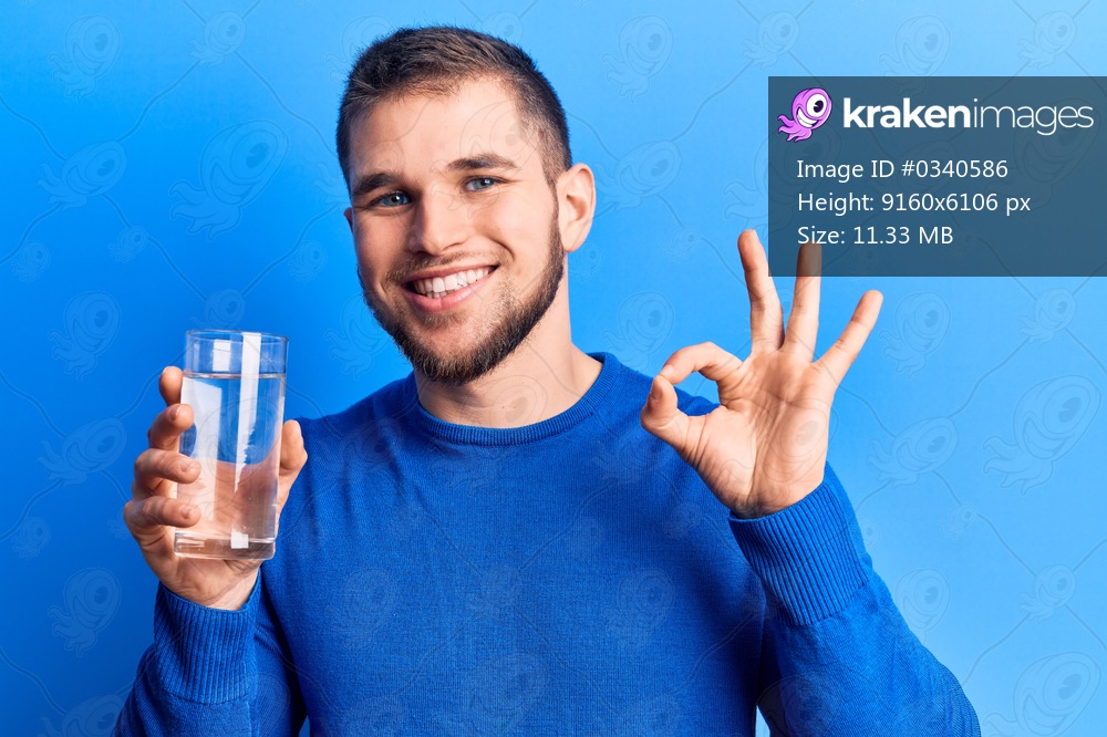 Young handsome man drinking glass of water doing ok sign with fingers, smiling friendly gesturing excellent symbol 