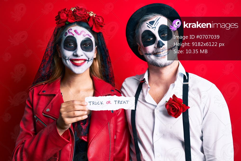Couple wearing day of the dead costume holding trick or treat paper looking positive and happy standing and smiling with a confident smile showing teeth 