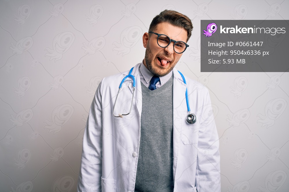 Young doctor man with blue eyes wearing medical coat and stethoscope over isolated background sticking tongue out happy with funny expression. Emotion concept.