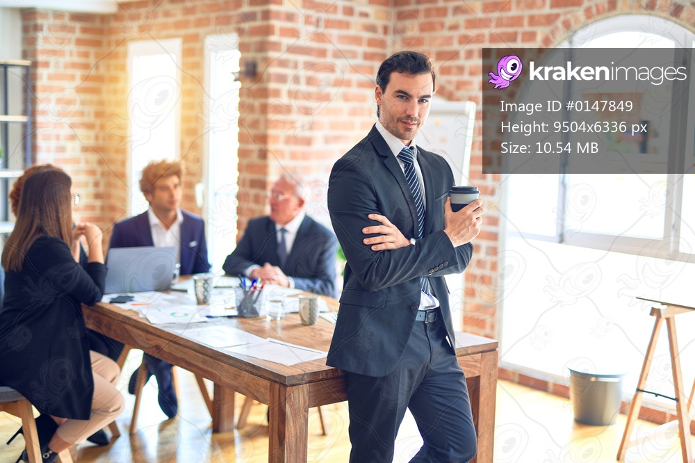 Group of business workers smiling happy and confident working together in a meeting. One of them, standing with smile on face looking at camera drinking coffee at the office.