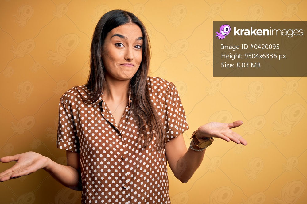 Young beautiful brunette woman wearing casual shirt over isolated yellow background clueless and confused expression with arms and hands raised. Doubt concept.