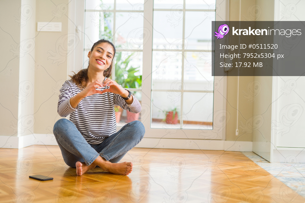 Young beautiful woman sitting on the floor at home smiling in love showing heart symbol and shape with hands. Romantic concept.