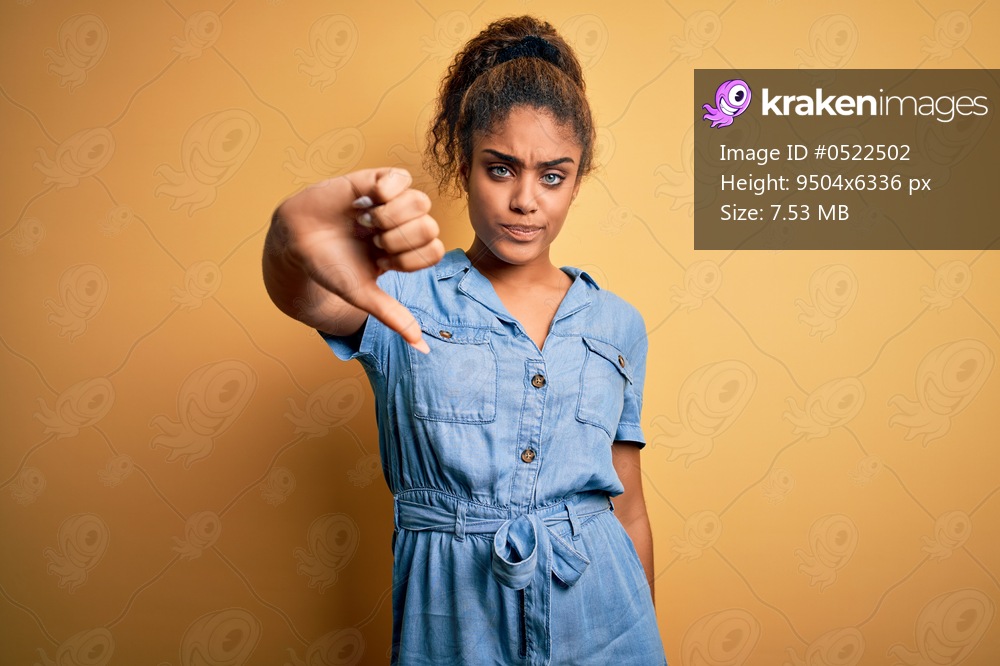 Young beautiful african american girl wearing denim dress standing over yellow background looking unhappy and angry showing rejection and negative with thumbs down gesture. Bad expression.
