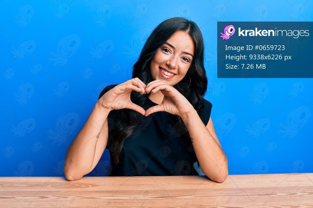 Beautiful brunette young woman wearing casual clothes sitting on the table smiling in love doing heart symbol shape with hands. romantic concept. 