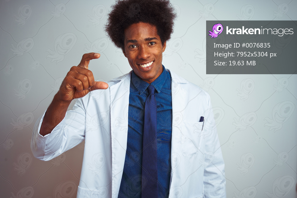 Young african american doctor man wearing coat standing over isolated white background smiling and confident gesturing with hand doing small size sign with fingers looking and the camera. Measure concept.