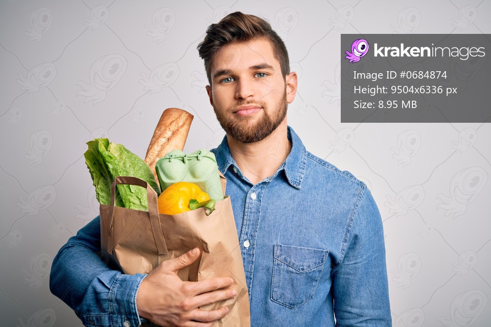 Young blond man with beard and blue eyes holding paper bag with food over white background with a confident expression on smart face thinking serious