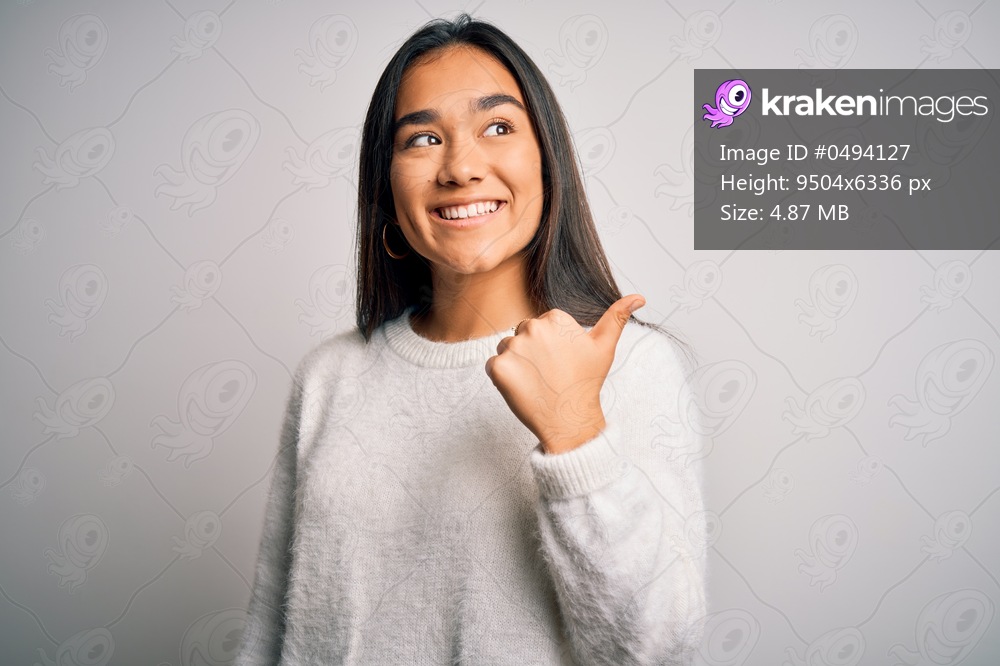 Young beautiful asian woman wearing casual sweater standing over white background smiling with happy face looking and pointing to the side with thumb up.