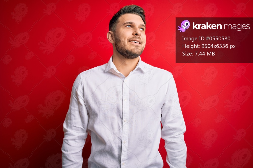 Young business man with blue eyes wearing elegant shirt standing over red isolated background looking away to side with smile on face, natural expression. Laughing confident.