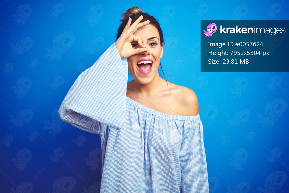 Young beautiful woman wearing bun hairstyle over blue isolated background doing ok gesture with hand smiling, eye looking through fingers with happy face.