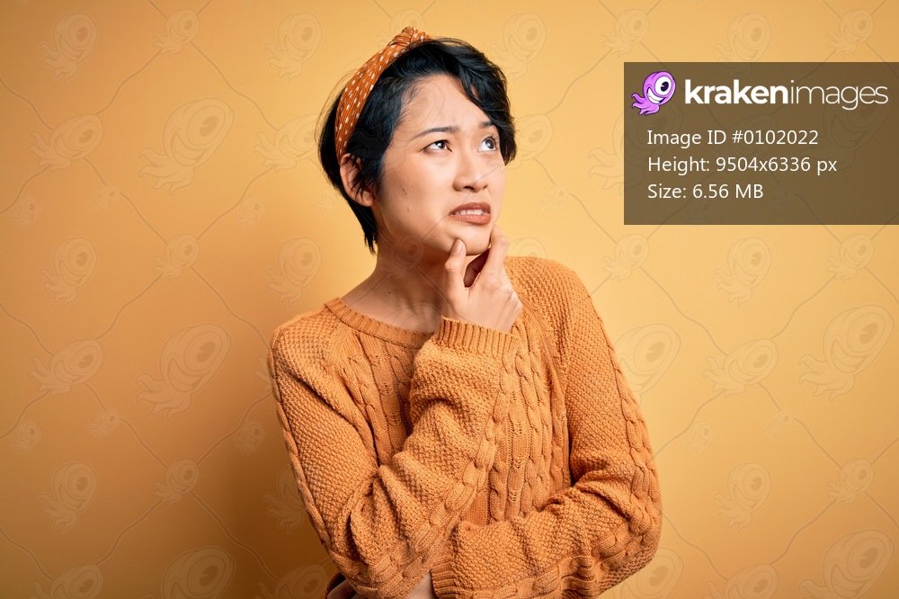Young beautiful asian girl wearing casual sweater and diadem standing over yellow background Thinking worried about a question, concerned and nervous with hand on chin