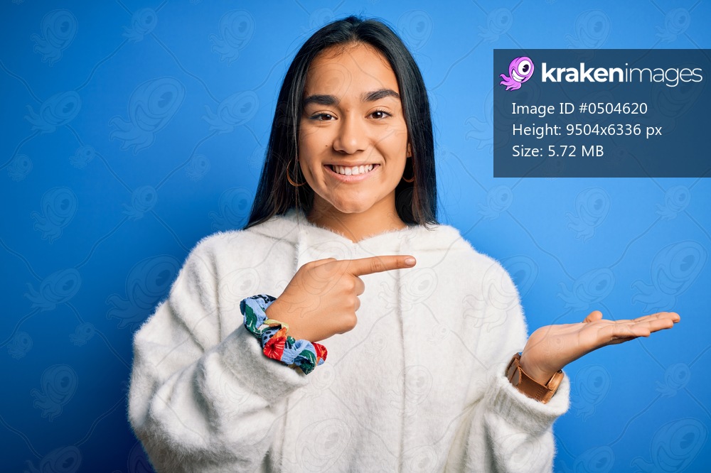 Young beautiful asian sportswoman wearing sweatshirt standing over isolated blue background amazed and smiling to the camera while presenting with hand and pointing with finger.
