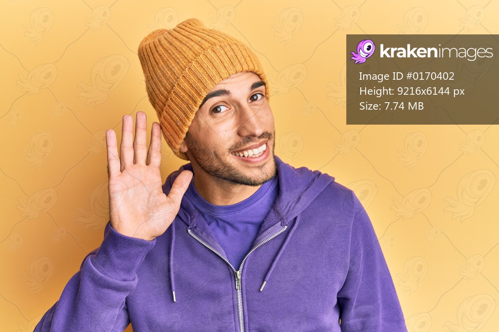 Young handsome man wearing wool winter hat smiling with hand over ear listening and hearing to rumor or gossip. deafness concept. 