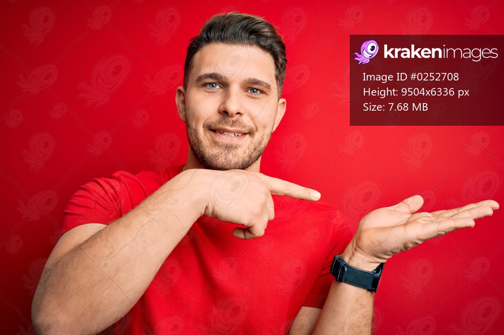 Young man with blue eyes wearing casual t-shirt over red isolated background amazed and smiling to the camera while presenting with hand and pointing with finger.