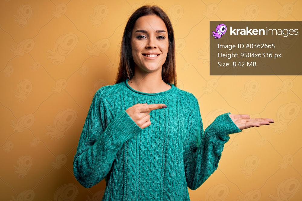 Young beautiful brunette woman wearing green casual sweater over yellow background amazed and smiling to the camera while presenting with hand and pointing with finger.