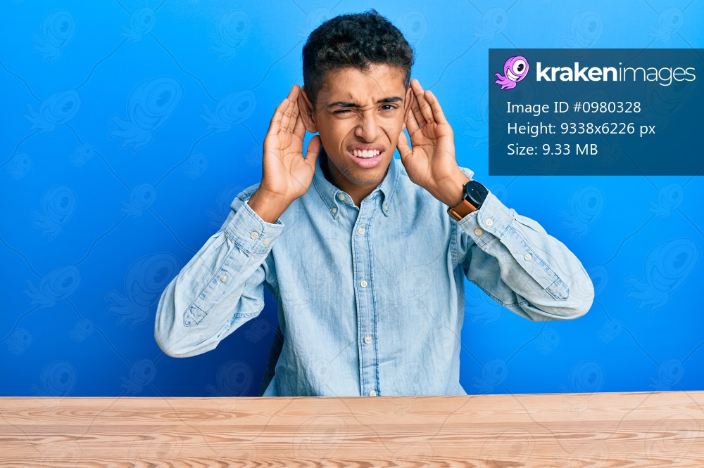 Young handsome african american man wearing casual clothes sitting on the table trying to hear both hands on ear gesture, curious for gossip. hearing problem, deaf 