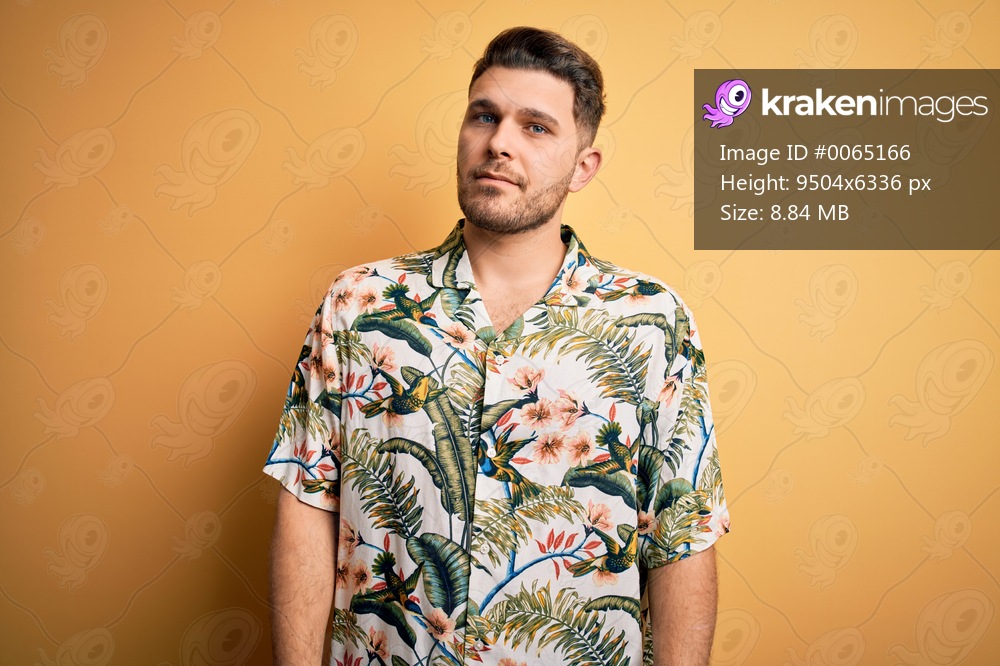 Young man with blue eyes on vacation wearing floral summer shirt over yellow background Relaxed with serious expression on face. Simple and natural looking at the camera.