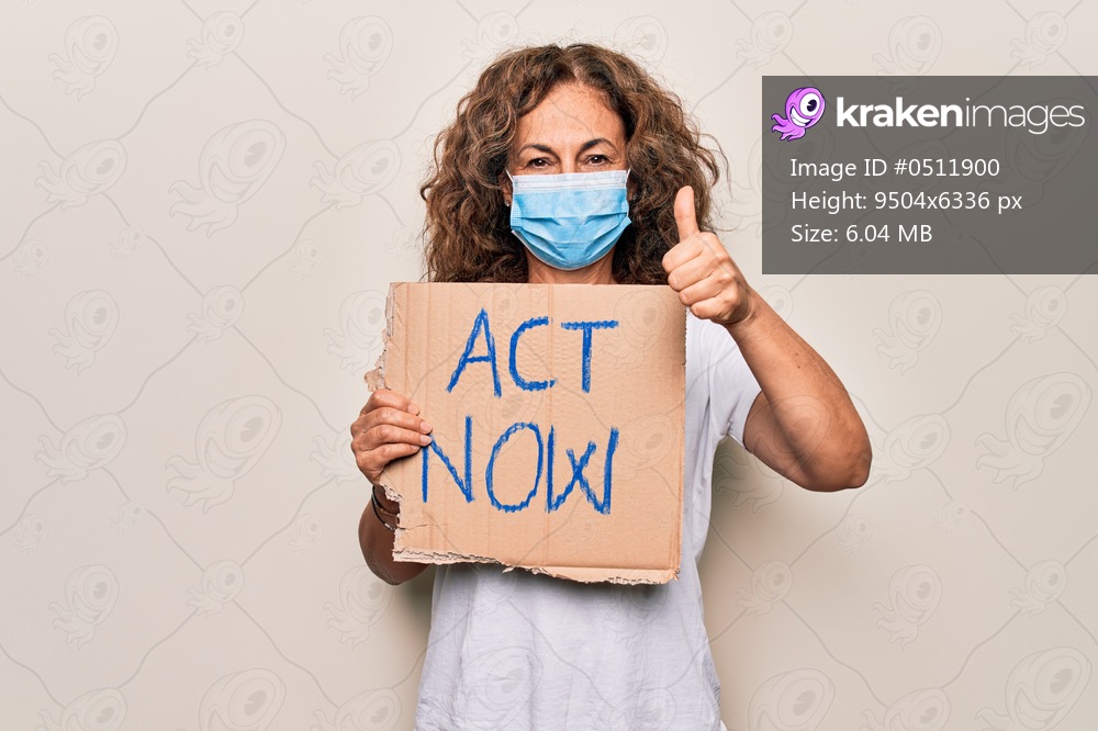 Middle age woman wearing coronavirus protection mask holding act now protest cardboard smiling happy and positive, thumb up doing excellent and approval sign