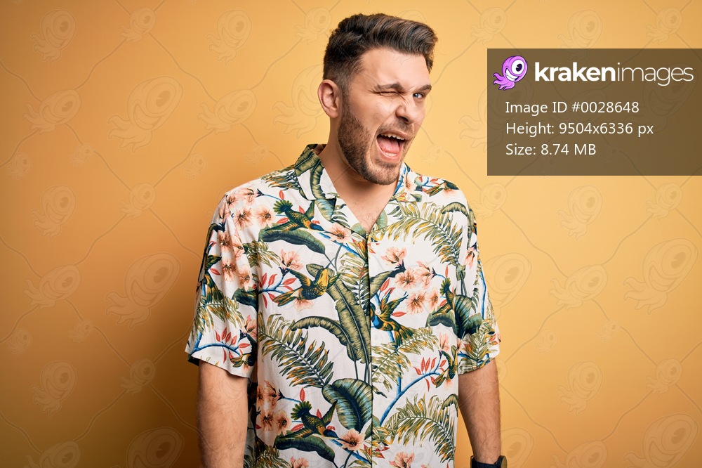 Young man with blue eyes on vacation wearing floral summer shirt over yellow background winking looking at the camera with sexy expression, cheerful and happy face.