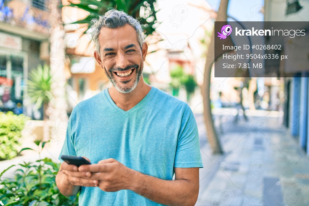 Middle age grey-haired man smiling happy using smartphone walking at street of city.