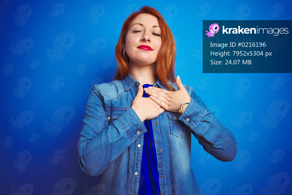 Young beautiful redhead woman wearing denim shirt standing over blue isolated background smiling with hands on chest with closed eyes and grateful gesture on face. Health concept.