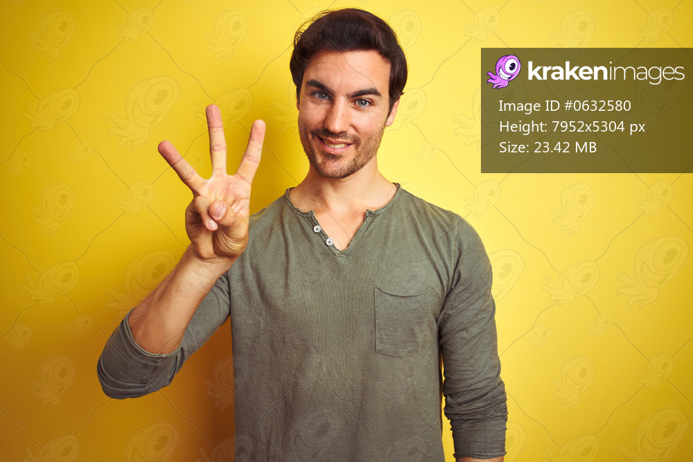 Young handsome man wearing casual t-shirt standing over isolated yellow background showing and pointing up with fingers number three while smiling confident and happy.