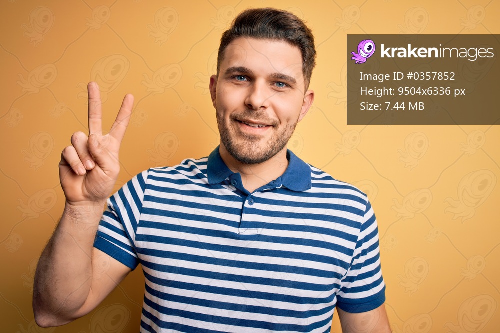 Young man with blue eyes wearing casual striped t-shirt over yellow background smiling with happy face winking at the camera doing victory sign. Number two.