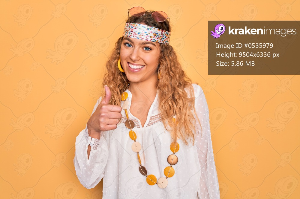 Young beautiful blonde hippie woman with blue eyes wearing sunglasses and accessories doing happy thumbs up gesture with hand. Approving expression looking at the camera showing success.
