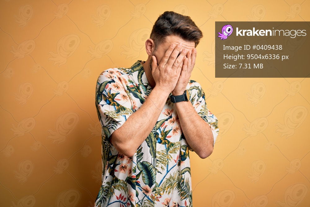 Young man with blue eyes on vacation wearing floral summer shirt over yellow background with sad expression covering face with hands while crying. Depression concept.