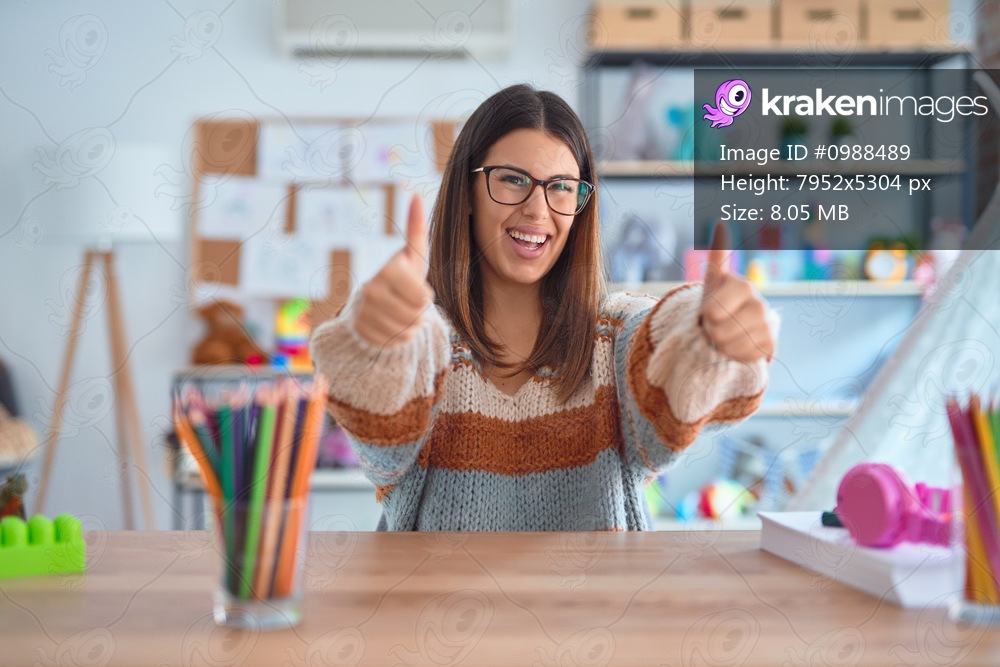Young beautiful teacher woman wearing sweater and glasses sitting on desk at kindergarten approving doing positive gesture with hand, thumbs up smiling and happy for success. Winner gesture.