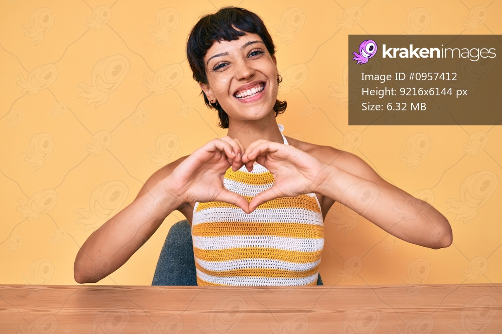 Beautiful brunettte woman sitting on the table over yellow background smiling in love showing heart symbol and shape with hands. romantic concept. 