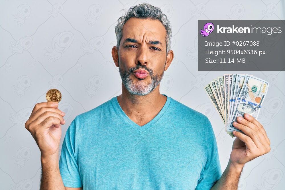 Middle age grey-haired man holding dollars and bitcoin looking at the camera blowing a kiss being lovely and sexy. love expression. 