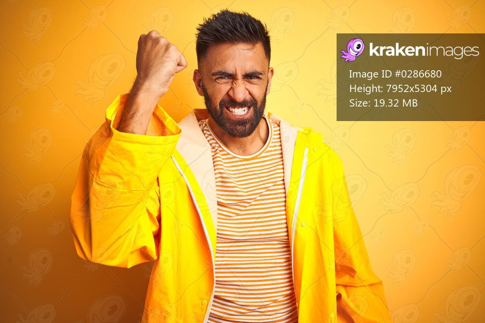 Young indian man wearing raincoat standing over isolated yellow background angry and mad raising fist frustrated and furious while shouting with anger. Rage and aggressive concept.