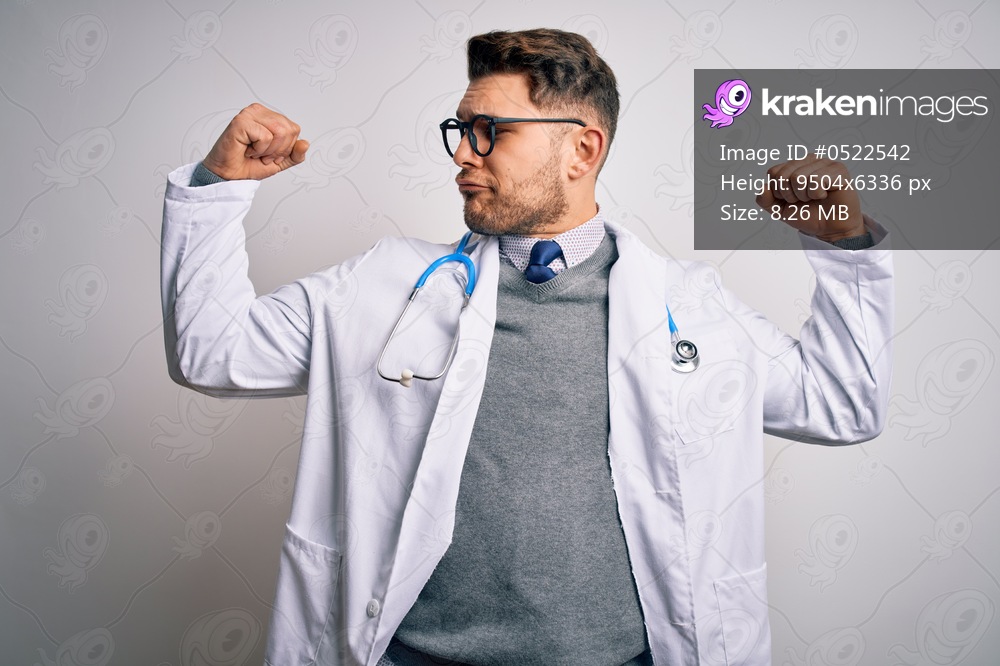 Young doctor man with blue eyes wearing medical coat and stethoscope over isolated background showing arms muscles smiling proud. Fitness concept.