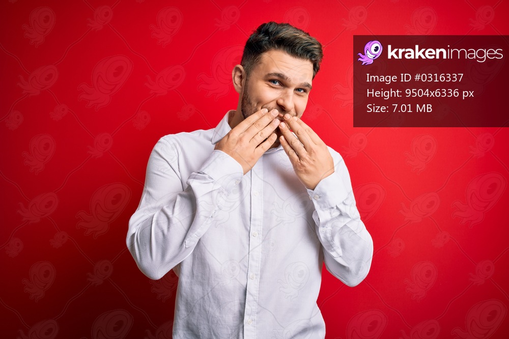 Young business man with blue eyes wearing elegant shirt standing over red isolated background laughing and embarrassed giggle covering mouth with hands, gossip and scandal concept