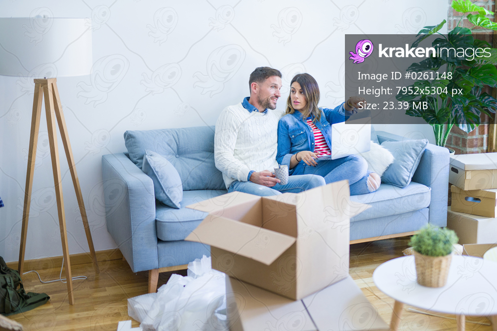 Young beautiful couple sitting on the sofa drinking cup of coffee using laptop at new home around cardboard boxes