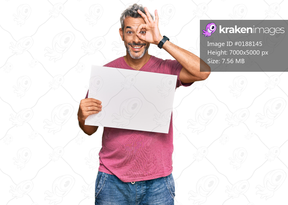 Middle age grey-haired man holding blank empty banner smiling happy doing ok sign with hand on eye looking through fingers 