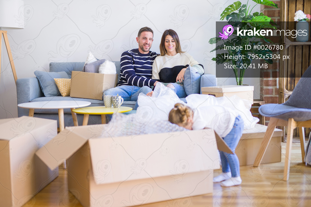 Beautiful family, parents sitting on the sofa drinking coffee looking his kid playing at new home around cardboard boxes
