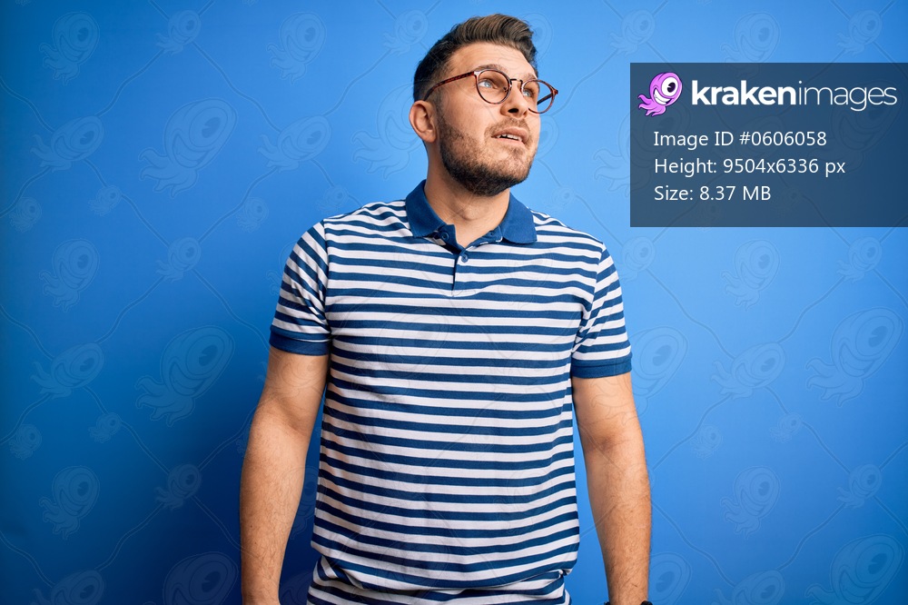 Young man with blue eyes wearing glasses and casual striped t-shirt over blue background looking away to side with smile on face, natural expression. Laughing confident.