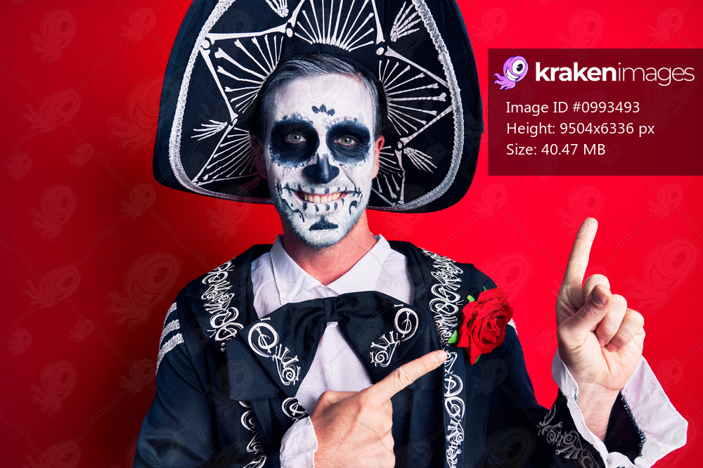 Young man wearing mexican day of the dead costume over red smiling and looking at the camera pointing with two hands and fingers to the side. 