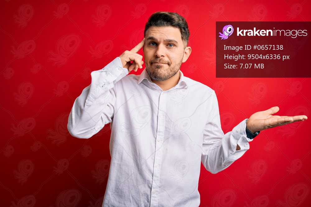 Young business man with blue eyes wearing elegant shirt standing over red isolated background confused and annoyed with open palm showing copy space and pointing finger to forehead. Think about it.