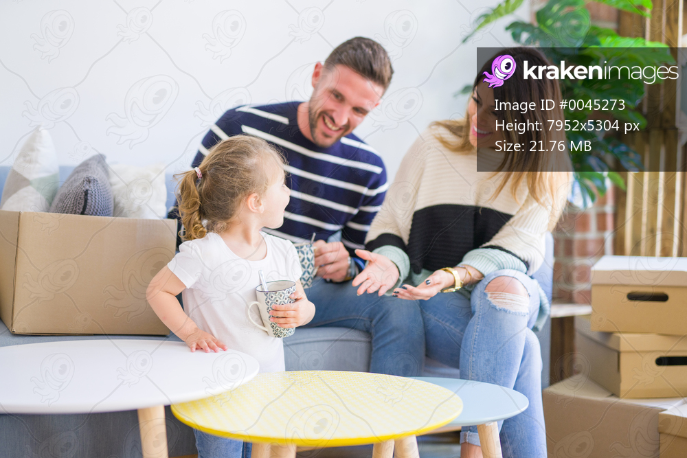 Beautiful family, parents sitting on the sofa drinking coffee looking his kid playing at new home around cardboard boxes