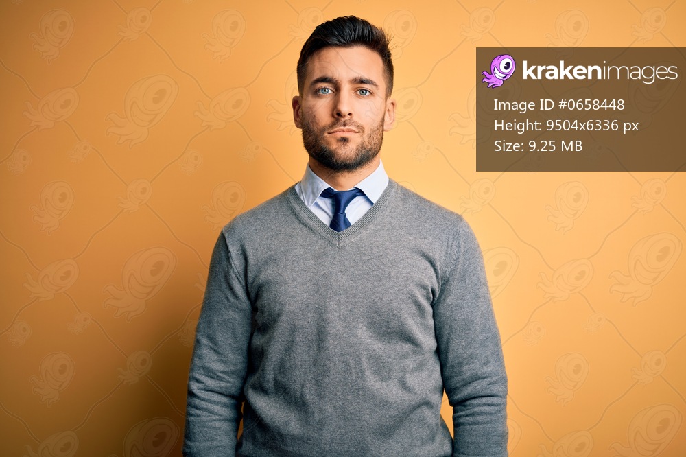 Young handsome businessman wearing elegant sweater and tie over yellow background with serious expression on face. Simple and natural looking at the camera.
