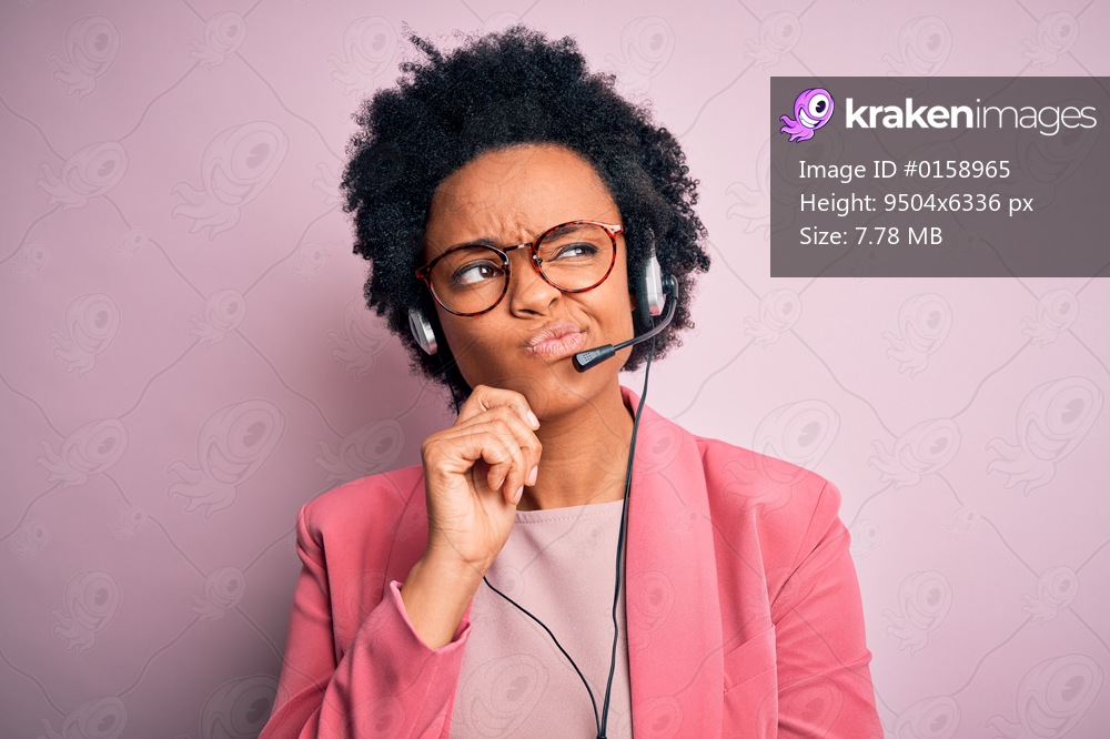 Young African American call center operator woman with curly hair using headset with hand on chin thinking about question, pensive expression. Smiling with thoughtful face. Doubt concept.