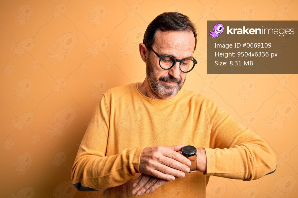 Middle age hoary man wearing casual sweater and glasses over isolated yellow background Checking the time on wrist watch, relaxed and confident