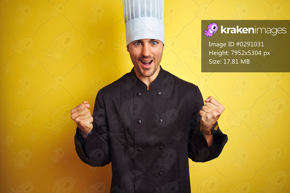 Young chef man wearing uniform and hat standing over isolated yellow background celebrating surprised and amazed for success with arms raised and open eyes. Winner concept.