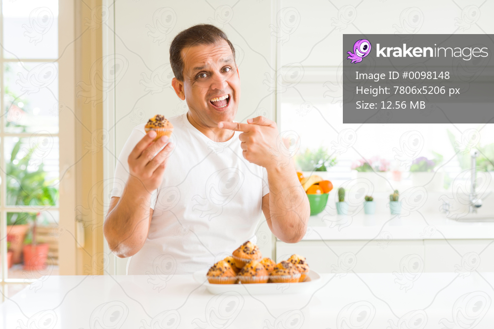 Middle age man eating chocolate chip muffins at home very happy pointing with hand and finger